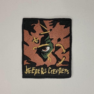 May include: A black and brown embroidered patch with a green eye peering out from a cracked wall. The text "Jeepers Creepers" is embroidered in yellow below the eye.
