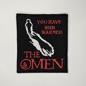 May include: Black felt patch with white embroidered image of a hand reaching out and red embroidered text that reads "You have been warned The Omen".