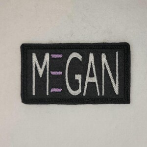 Puede incluir: Parche bordado negro con letras blancas que dicen "MEGAN" con costuras moradas.