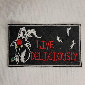 Puede incluir: Parche bordado en blanco y negro con una cabeza de cabra y el texto "LIVE DELICIOUSLY" en rojo.
