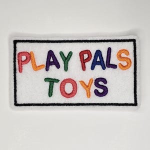 Puede incluir: Un parche rectangular blanco con borde negro, que presenta el texto "PLAY PALS TOYS" en letras bordadas de colores.