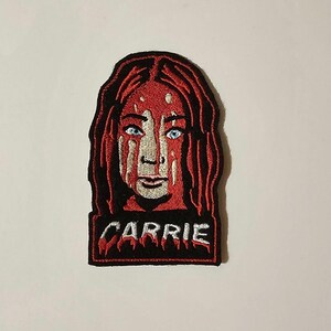 May include: Embroidered patch featuring a red-haired woman with blood dripping down her face. The patch has a black background with the word "CARRIE" in white lettering.