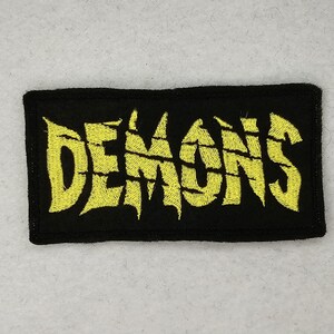 May include: Black patch with yellow embroidered text that reads "DEMONS" in a jagged font.
