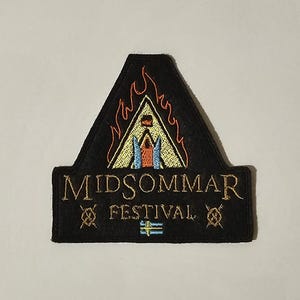 May include: Black embroidered patch with the text "MIDSOMMAR FESTIVAL". The patch features a triangular design with flames, a central structure, and the Swedish flag. The patch is a unique souvenir.