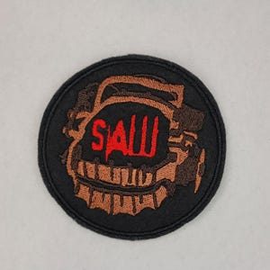May include: A black and brown embroidered patch with the word "SAW" in red lettering. The patch features a circular design with a stylized image of a trap.