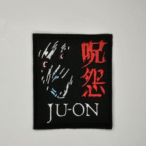 May include: Black square patch with embroidered design. Features a stylized face in white and blue on the left, and red Japanese characters on the right. The word "JU-ON" is embroidered in white at the bottom.