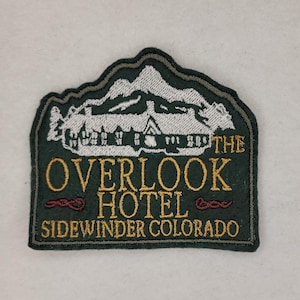 Peut inclure: Patch brodé vert avec du texte blanc qui dit "The Overlook Hotel Sidewinder Colorado". Le patch présente une silhouette blanche d'un bâtiment avec une montagne derrière.