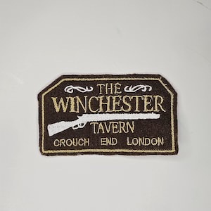 Può includere: Una toppa di feltro marrone con testo ricamato in oro che recita "The Winchester Tavern Crouch End London". Un fucile ricamato in bianco è sopra il testo.