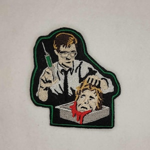 May include: Embroidered iron-on patch featuring a man in a white shirt and glasses holding a syringe and a severed head in a box. The man has a serious expression on his face. The patch is green, black, and red.