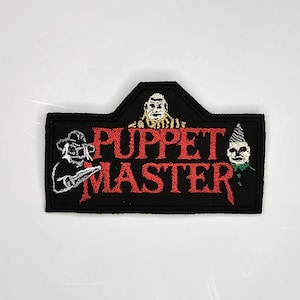 May include: Black embroidered patch with the words "Puppet Master" in red. The patch features three puppet characters with white and black details.
