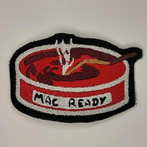 Puede incluir: Un parche bordado rojo y blanco con un borde negro. El parche representa un tazón rojo con un remolino marrón en el interior y un palo amarillo que sobresale. El texto "MAC READY" está escrito en blanco en la parte inferior del tazón.