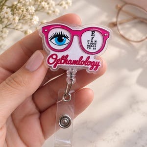 May include: A clear acrylic badge reel featuring a pink glasses frame design. The left lens shows a blue eye with long lashes, and the right lens displays the text E F CAN P YOU READ? TH IS. The word Opthamlology is printed in pink script below.