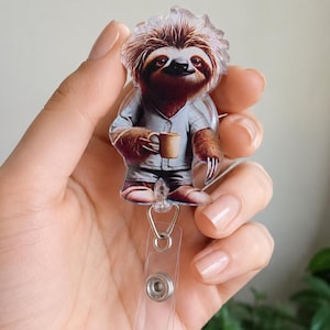 May include: An acrylic badge reel featuring a brown sloth with messy fur wearing light gray pajamas and holding a tan coffee mug. The sloth is printed on a clear acrylic shape attached to a retractable cord mechanism with a clear plastic strap.