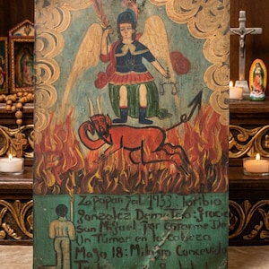 May include: A rectangular retablo painting on wood depicting a winged figure in blue and green robes standing over a red horned devil amidst orange flames. The lower section features a kneeling figure and Spanish text: Zapopan Jal 1953.
