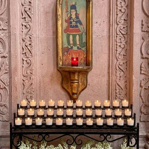 May include: A black metal votive candle stand with three rows of lit candles in clear glass holders, positioned below a gold-framed wall niche containing a painted figure of a saint standing over flames, with three terracotta vases of white flowers.