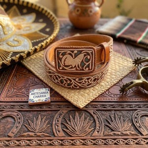 May include: A tan leather belt featuring an intricate floral pattern and a rectangular buckle with a carved rooster design. A small square tile with the text ARTESANÍA CHARRA is placed next to the belt, which is coiled on a woven mat.