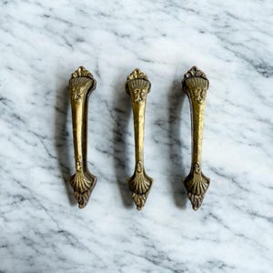May include: Three antique brass drawer pulls with ornate detailing. Each pull features a decorative top with a face design, a slender handle, and a stylized base. The pulls are arranged on a marble surface.