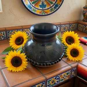 Puede incluir: Un jarrón de cerámica negro con diseños grabados y una base con cuentas, rodeado de girasoles amarillos brillantes. El jarrón se exhibe sobre una superficie de baldosas con acentos decorativos coloridos, creando una exhibición rústica y artística.