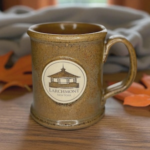 Puede incluir: Una taza de cerámica marrón con asa, con un emblema circular blanco con una ilustración de un mirador y el texto "LARCHMONT NEW YORK". La taza tiene una superficie texturizada y un aspecto rústico.