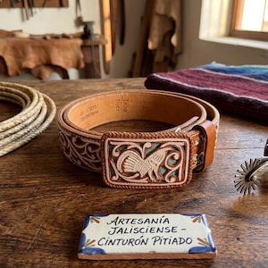 May include: A tan leather belt with intricate hand-tooled floral patterns and a rectangular buckle featuring a carved rooster design. A small rectangular tile in front displays the text Artesanía Jalisciense - Cinturón Pitiado in black ink.
