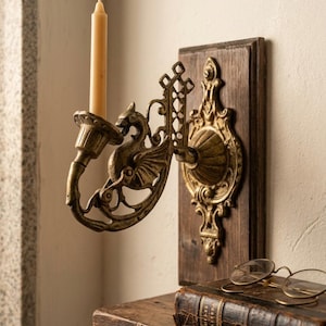 Puede incluir: Un candelabro de pared con una figura de dragón de metal dorado fijada a una placa rectangular de madera oscura. El soporte sostiene una vela cónica de color crema. Debajo, un libro antiguo encuadernado en cuero tiene unas gafas de metal.