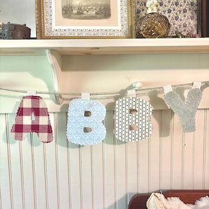May include: A decorative banner spelling "BABY" with fabric letters. The letter "A" is red and white gingham, "B" is light blue with a floral pattern, "B" is navy with white dots, and "Y" is gray. The banner is strung on a white cord.