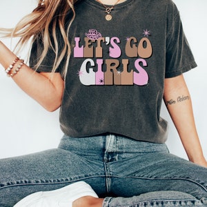 May include: A dark gray t-shirt with a retro-style graphic that says "Let's Go Girls" in pink, brown, and white letters. The graphic includes a small pink cowboy hat.