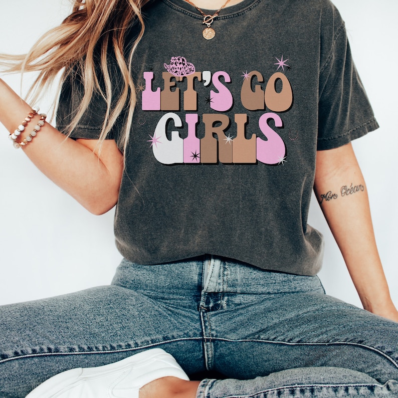 May include: A dark gray t-shirt with a retro-style graphic that says "Let's Go Girls" in pink, brown, and white letters. The graphic includes a small pink cowboy hat.