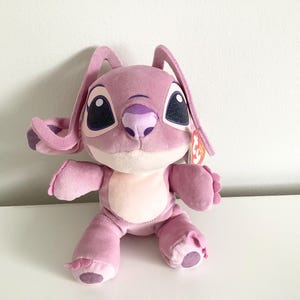 May include: A plush purple Stitch character with large eyes and a pink nose. The character is wearing a pink and purple beanie and has a Ty tag attached to its ear.