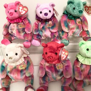 Op de afbeelding: Een verzameling van zes kleurrijke pluche teddyberen. Elke beer heeft een andere kleur kop en een gefranjerde kraag. De beren hebben een tie-dye patroon op hun lichaam en de maand van het jaar staat op een label.