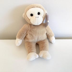 May include: A beige plush monkey toy with black eyes and a white face. The monkey is sitting on a white surface.