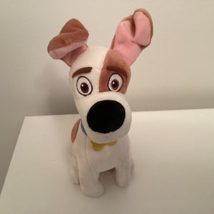 Op de afbeelding: Een pluche hondenspeelgoed met witte vacht, bruine oren en een bruine vlek over één oog. De hond heeft een zwarte neus, bruine ogen en een blauwe halsband met een gele tag. Het speelgoed staat op een wit oppervlak.