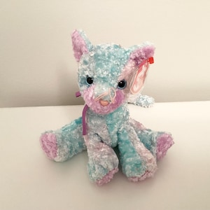 May include: A blue and pink plush cat with a purple ribbon around its neck. The cat has a white face with pink nose and whiskers. The cat is sitting on a white surface.