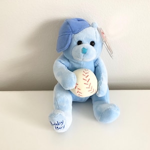 May include: A blue plush toy bear wearing a blue baseball cap and holding a white baseball with red stitching. The bear has a blue nose and blue eyes. The bear's foot has the words "Baby Boy" embroidered on it.