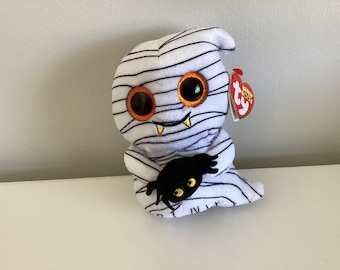 May include: A white plush mummy toy with black stripes, orange and black eyes, and small yellow fangs. It holds a black spider with yellow eyes. A red heart-shaped tag is attached. Halloween-themed plush toy.