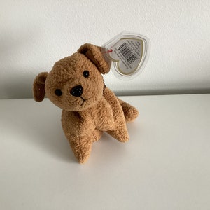 Könnte beinhalten: Ein braunes und beiges Plüsch-Hundespielzeug mit schwarzer Nase und Augen. Das Spielzeug hat ein herzförmiges Etikett mit dem Text "Ty Inc. Beanie Babies."