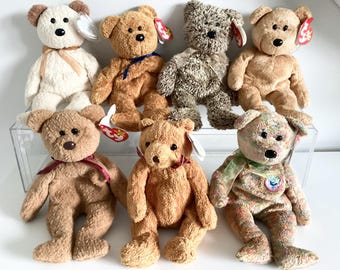 May include: Collection of plush teddy bears in various colors, including cream, brown, and multi-colored. Each bear has a distinct design, with some featuring ribbons and tags. The bears are displayed on a clear shelf.