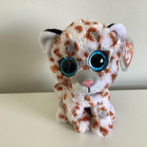 May include: A white and brown plush toy leopard with blue eyes and a red nose. The toy is wearing a clear plastic pair of glasses and has a red and white Ty Beanie Baby tag attached to its ear.