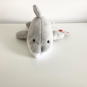 May include: A grey plush shark toy with black eyes and a white mouth. The toy has a red tag attached to it.