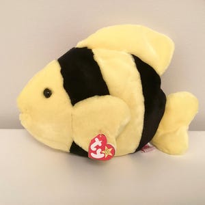 May include: A soft toy fish in yellow and black stripes, with a yellow fin and tail. The stuffed animal has a red heart-shaped tag with a gold star. The toy is designed to resemble a tropical fish.