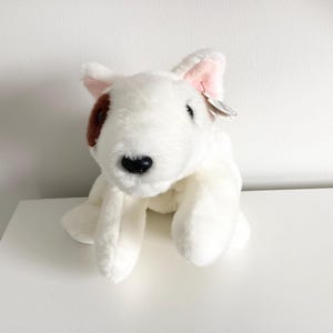 Peut inclure: Peluche représentant un chien Bull Terrier blanc, avec une tache marron sur un œil et l'intérieur des oreilles rose. Le jouet a un nez noir et est posé sur une surface blanche. Une petite étiquette est attachée à l'oreille.