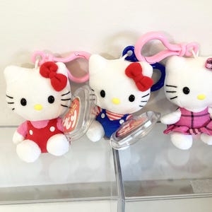 May include: Three plush Hello Kitty keyrings. Each white cat has a red or pink bow, black eyes, and a yellow nose. One wears a red polka-dot dress, another a blue outfit, and the third a pink plaid dress. Each has a plastic clip.
