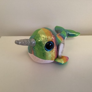 May include: A plush toy narwhal with a rainbow iridescent finish and a silver glitter horn. The narwhal has a single blue eye and a white belly.
