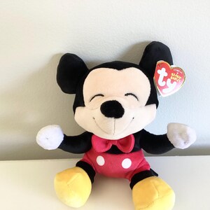 May include: A plush Mickey Mouse toy with black ears, a black nose, and a smiling face. The toy has a red bow tie, red shorts with white buttons, white gloves, and yellow shoes. A Ty Beanie Babies tag is attached.