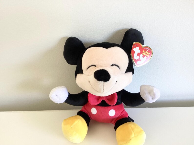 May include: A plush Mickey Mouse toy with black ears, a black nose, and a smiling face. The toy has a red bow tie, red shorts with white buttons, white gloves, and yellow shoes. A Ty Beanie Babies tag is attached.