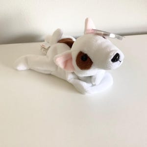 May include: A white plush toy dog with brown spots around one eye and on its back. The dog has pink inner ears and a black nose. A clear tag is attached to the dog's ear.