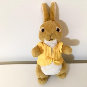 May include: A plush toy rabbit with tan fur, wearing a yellow vest with a bow. The rabbit has brown eyes and a white chest. The toy is standing upright against a white background.