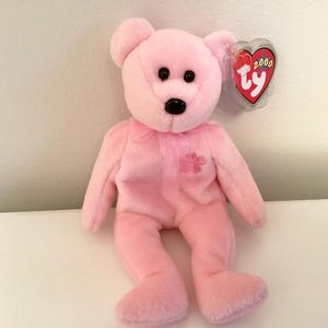 May include: A pink plush teddy bear with black eyes and a black nose. The bear has a pink ribbon around its neck and a small flower on its chest. A red heart-shaped tag with the word "Ty" is attached.