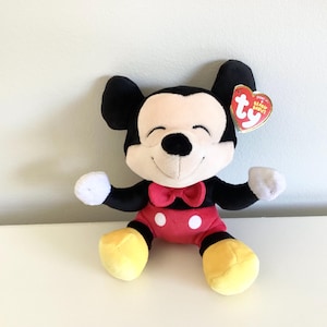 May include: A plush Mickey Mouse toy with black ears, arms, and a smiling face. He wears red shorts with white buttons, a red bow tie, and yellow shoes. A Ty Beanie Babies tag is attached.
