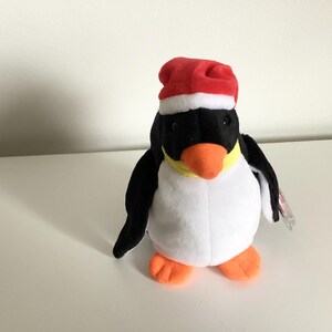 Op de afbeelding: Een pluche pinguïn met een kerstmuts. De pinguïn heeft een zwart lichaam, een witte buik, oranje voeten en snavel en een gele nek. De rode kerstmuts heeft een witte rand. Er is een klein label bevestigd.